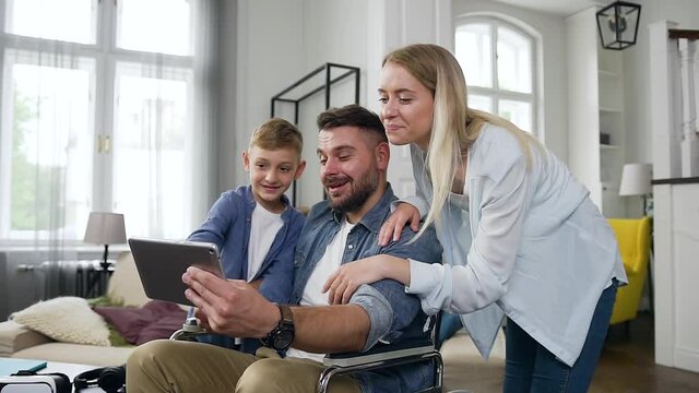 Good-tempered positive smiling boy,woman and man in wheelchair talking on applying i-pad in contemporary living-room