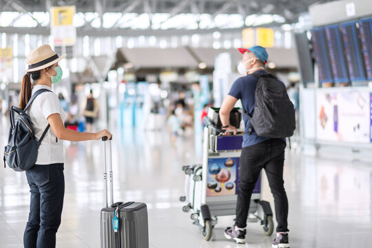 Young female wearing face mask with luggage checking flight time in airport, protection Coronavirus disease (Covid-19) infection, Asian woman traveler with hat. New Normal and travel bubble concept