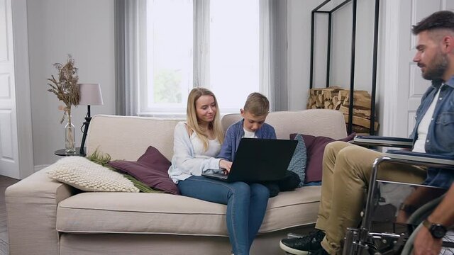 Front view of attractive contented friendly mother which using computer together with her interested smart 8-aged son on the couch when handsome bearded disabled father in wheelchair joining to them