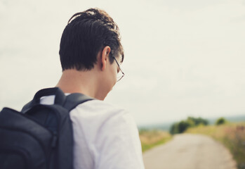 A teenager in glasses and a white t-shirt and a backpack goes to school to study.Country nature. Back to school.