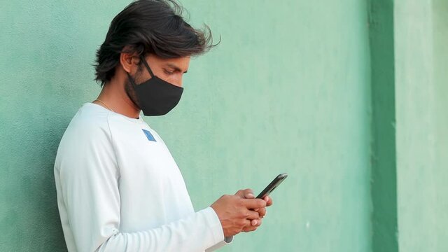 Young man in medical mask busy in using mobile phone during workout at outdoors - Concept of netizen smartphone addiction, protection from coronavirus or covid-19.