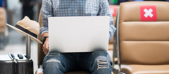 Young male wearing face mask and using computer laptop in airport, protection Coronavirus disease infection, Asian man traveler sitting on chair. New Normal, social distancing and digital nomad