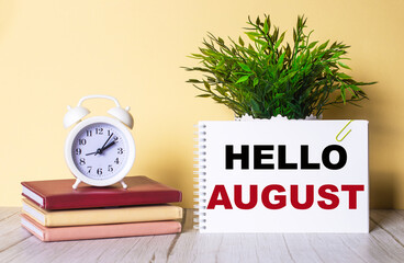 Hello August written on a white card next to a white alarm clock and diaries. Communication concept.