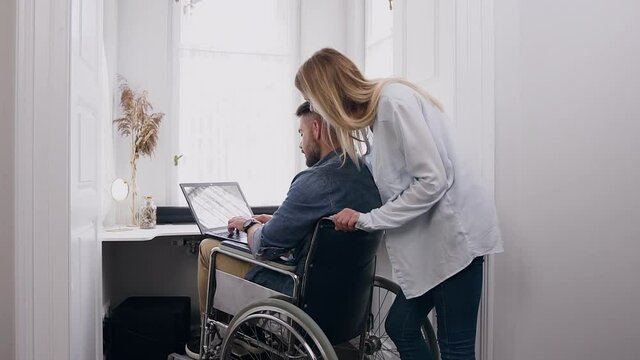 Handsome Happy Smiling Confident Young Bearded Man In Wheelchair Working On Laptop At Home And Secretly Looking At His Wife Which Standing Behind His Back