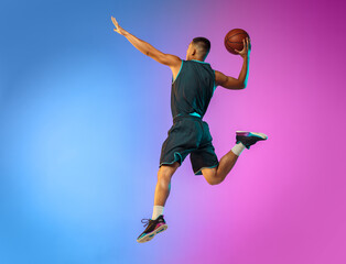 Fototapeta premium Winner. Young basketball player in action, motion in high jump on gradient background in neon. Concept of sport, movement, energy and dynamic, healthy lifestyle. Training, practicing, trendy colors.
