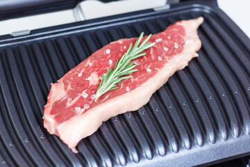 A piece of fresh meat with coarse salt and a sprig of rosemary. Juicy beef steak on the electric grill. - 368849874