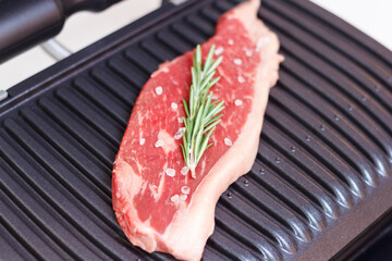 A piece of fresh meat with coarse salt and a sprig of rosemary. Juicy beef steak on the electric grill. - 368850250