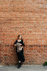 Lonesome young woman in a long black dress in front of a dirty brick house wall