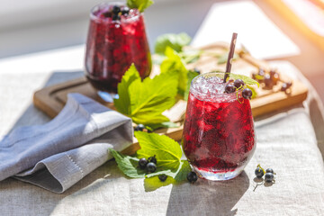 Two glass of cold ice black currant juice with ripe berries and green leaves on table in sunny room...