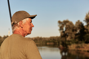 Side view of senior man sitting on bank of river and looking aside, having rest in open air, enjoying beautiful nature near lake, wearing green t shirt and cap.