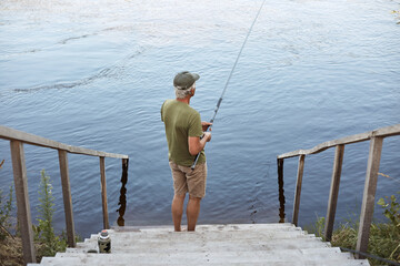 Fototapeta na wymiar Backwards view of man catching fish while posing on wooden stairs leading to lake, male wearing casual attire, enjoying beautiful nature and fishing.
