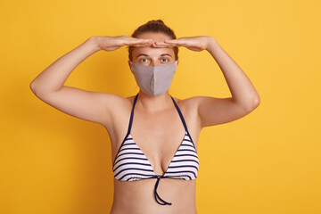 Winsome woman in bathing suit and medical mask poses against yellow background, keeping both palms near forehead, looking in distance, wears protective mask during quarantine.