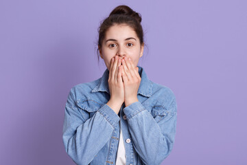Surprised young woman covering her mouth and looking directly at camera, wearing denim jacket, has dark hair and knot, posing isolated over lilac background.