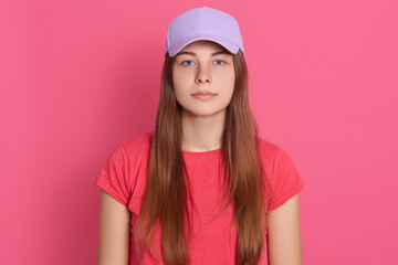 Beautiful woman wearing baseball cap and t shirt posing isolated over pink background, looks at camera with serious expression on face, having long straight hair.