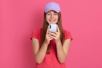 Satisfied lovely woman holding modern cell phone, texts with her friend online, dresses red t shirt and baseball cap, poses against pink wall, standing with charming smile.