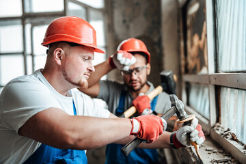 Two workers at a very big construction site removing plaster from an old brick wall near a glass window