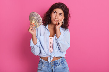 Young attractive smiling woman thinking how to spend her bunch of money, female wearing blue shirt and jeans, standing against pink wall with pensive expression.
