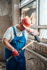 A tired worker in a special uniform stands at a construction site in a protective helmetand with a big sledgehammer in hand