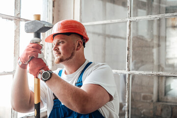 Worker, looking out the window, stands at the construction site. He is wearing a red safety helmet and he holds a sledgehammer for repairs in his hands.