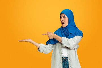 Pointing, smiling. Young muslim woman isolated on yellow studio background. Stylish, trendy and beautiful female model. Human emotions, facial expression, sales, ad, shopping concept. Copyspace.