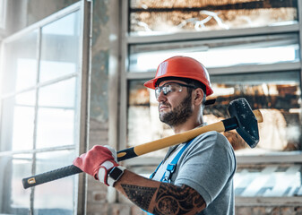 Attractive constructional worker with tattos stands near the window at a construction site. He is...