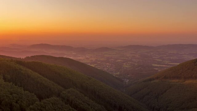 Timelapse of the sunset on the hillside and mountainous landscape below which lie several smaller villages the sun changes color from orange to dark blue during the west of the Beskydy region.