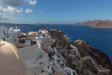 Santorini, Oia - View of white houses on a cliff above the Caldera, winding stairs leading to the sea. Beautiful blue sky with white clouds.