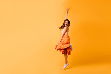Studio shot of pretty girl in orange skirt and white shoes. Excited red-haired lady jumping on...