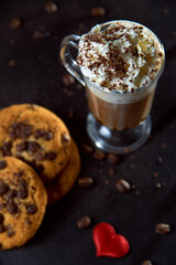 Close up of a glass cup of coffee with whipped cream and chocolate on it, chocolate chip cookies and roasted coffee beans on dark background. Concept of ready to eat food, tasty snack. Selective focus