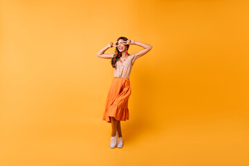 Full-length portrait of trendy emotional girl dancing on bright background. Jocund lady in long orange skirt expressing happiness.