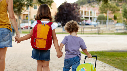 Mother and pupil and kids holding hands going to school in first class with schoolbag