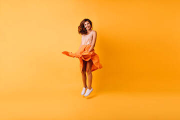 Fototapeta na wymiar Full-length portrait of debonair stylish lady jumping in studio. Gorgeous ginger girl in orange skirt having fun.