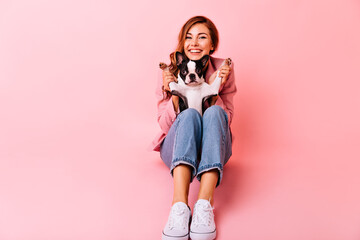 Well-dressed girl playing with french bulldog. Indoor photo of beautiful red-haired woman expressing sincere emotions.