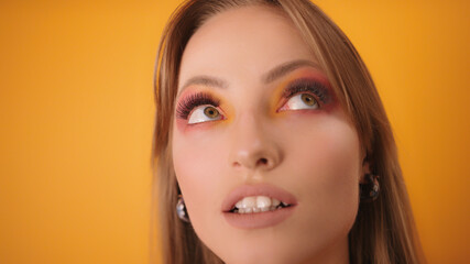 Fototapeta premium Portrait of Caucasian young woman with eye shadows and eyelashes extension looking surprised around. Isolated on the orange background. High quality photo
