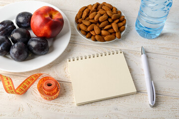 Paper notepad, measuring tape, fresh fruits, almonds and drinking water on a white wooden table. Copy space on notebook page. Concept of healthy eating, dieting, body slim.