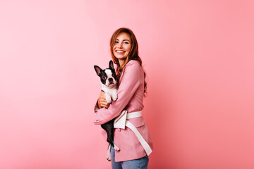 Pleasant caucasian woman in pink attire embracing puppy. Glamorous white girl holding french bulldog with smile.