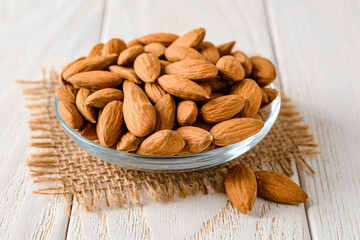 Tasty raw almonds in a glass saucer on a white wood table. Unroasted nuts as antioxidant and protein source for ketogenic diet and vegetarianism. Healthy eating.
