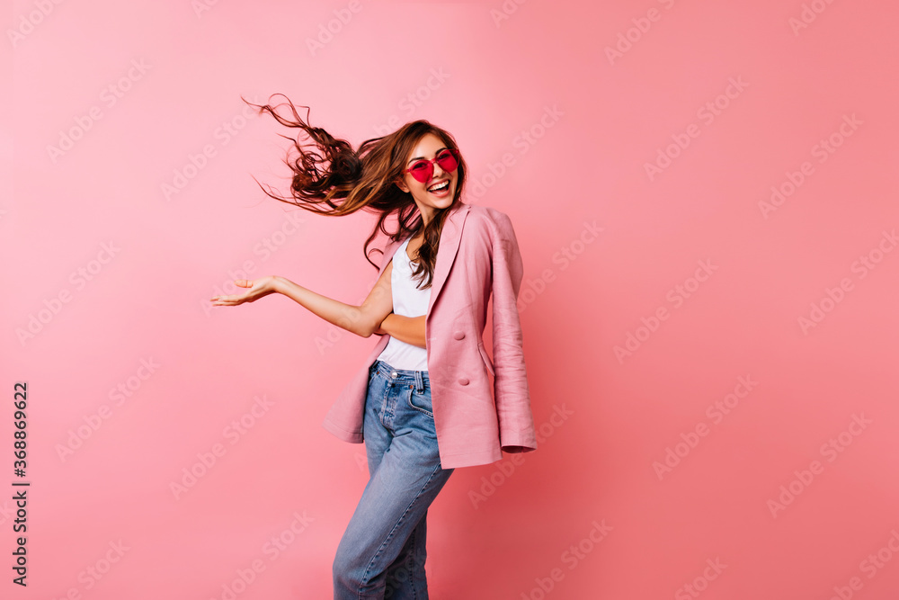 Wall mural Excited good-looking girl in sunglasses dancing in studio. Ginger blissful woman posing on rosy background with hair waving.