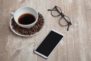wooden office table with a porcelain cup of coffee beans, reading glasses and a phone