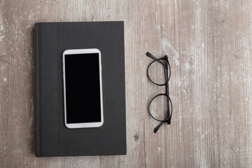 cell phone on a black book next to some classic glasses, on an old wooden table