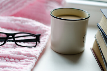 White cup with coffee, books, glasses and pink knitted scarf by the window. Autumn still life.