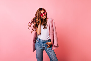 Excited white girl in bright stylish glasses posing on pink background. Dreamy curly woman playing with her ginger hair and laughing.