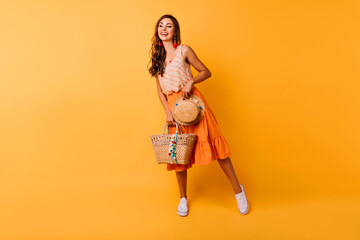 Full-length shot of inspired woman with summer accessories. Happy ginger female model in orange skirt holding hat and bag.