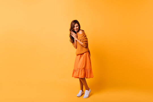 Full-length photo of cute young woman in white gumshoes isolated on yellow background. Studio portrait of gorgeous dark-haired girl in orange outfit.