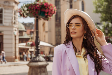 Outdoor fashion portrait of elegant woman wearing lilac suit, white hat, yellow sunglasses, posing in street of European city. Copy, empty space for text