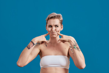 Portrait of a young half naked tattooed woman looking playful, having fun, holding air in her cheeks and pointing on them with fingers isolated over blue background