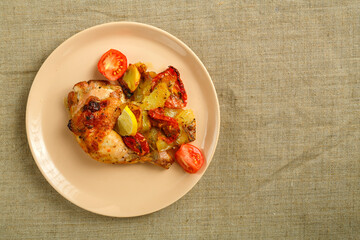 Baked chicken leg with potatoes and tomato on a beige plate on a linen tablecloth.
