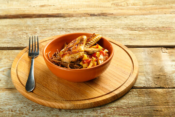 Brown cup with soba noodles with vegetables and chicken wings on a wooden stand and a fork.