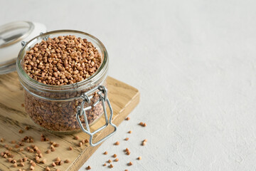 Buckwheat uncooked grains in glass jar on the wooden cutting board on gray background. Healthy, dietary food. Organic, carbohydrate product. Food storage. Close up, place for text.