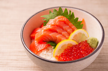 raw pink salmon sashimi and red salmon roe ikura over white rice with lemon wedges in a ceramic bowl